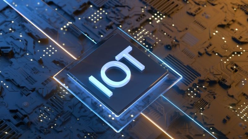 Imagem de um microchip com a sigla "IOT" em destaque, cercado por luzes e circuitos digitais, representando um dispositivo IoT para soluções industriais e proteção de máquinas e sistemas operacionais.